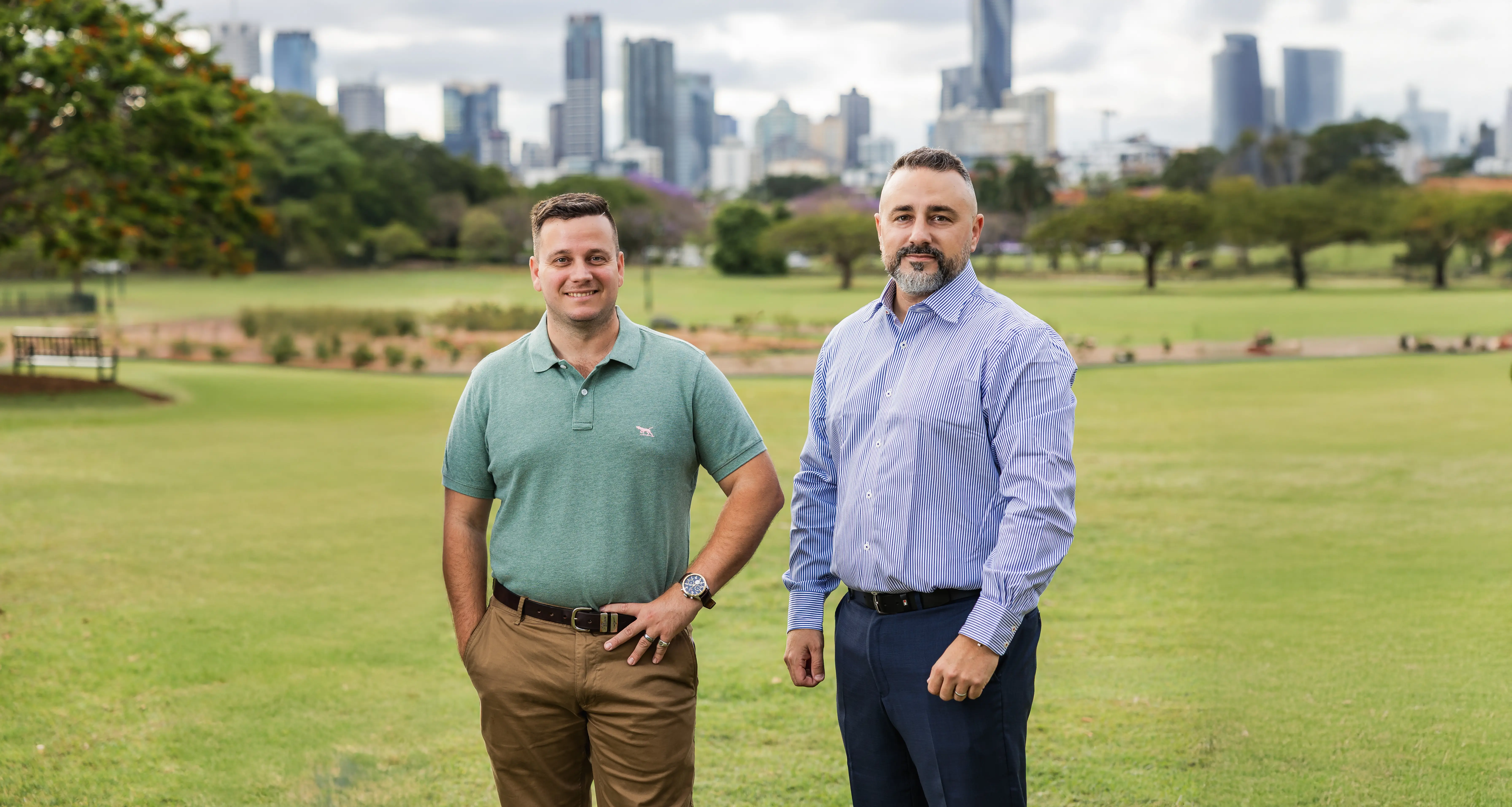 Two men standing on a green lawn with a city skyline blurred in the background, one wearing a green polo and brown pants and the other in a blue striped shirt and dark pants.
