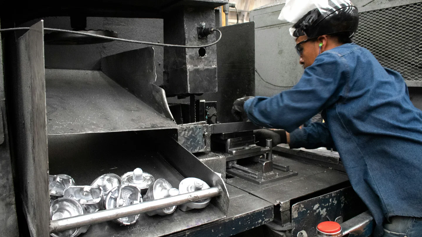 Factory worker wearing safety gear operates an industrial CNC metal cutting machine, with freshly cut metal components collected on a chute in the foreground.