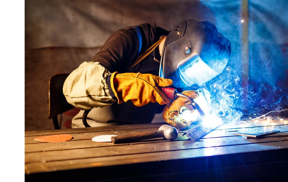 Welder wearing protective gloves and helmet using a welding torch on a metal piece, with sparks and smoke visible.