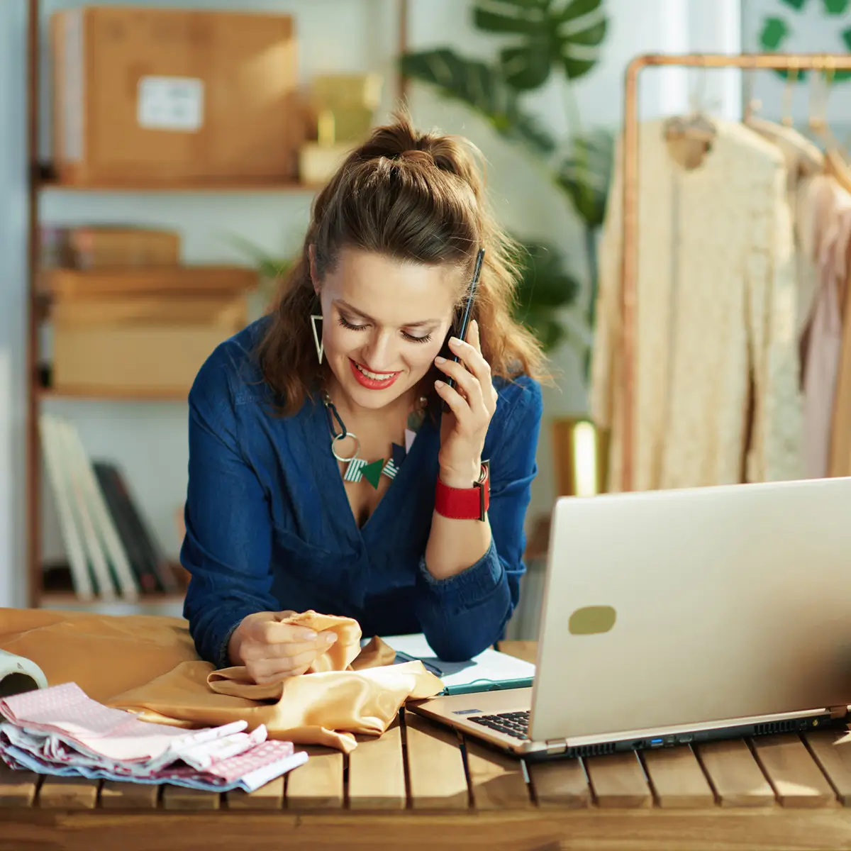 Smiling woman in blue shirt examining fabric while talking on a smartphone at a wooden table with a laptop and folded textiles.