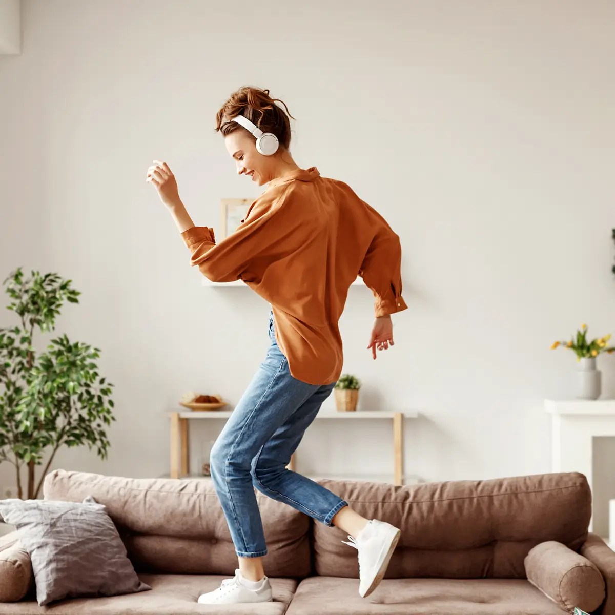 Smiling young woman dancing on a couch wearing headphones and casual clothes in a bright living room.