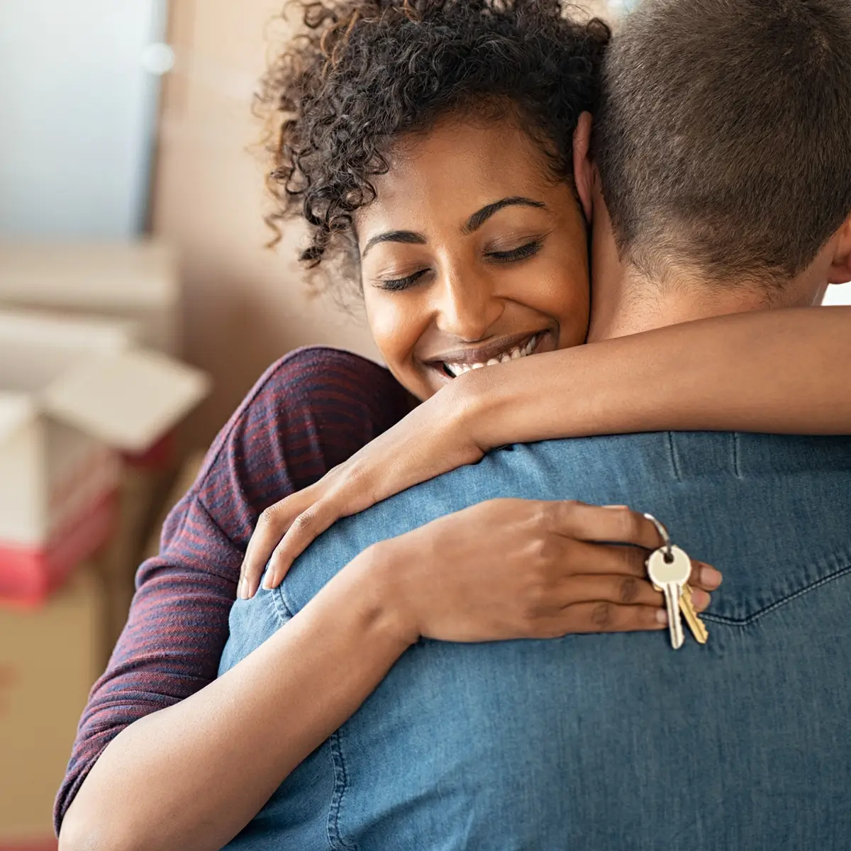 Happy woman hugging a man while holding house keys in a moving box-filled room.