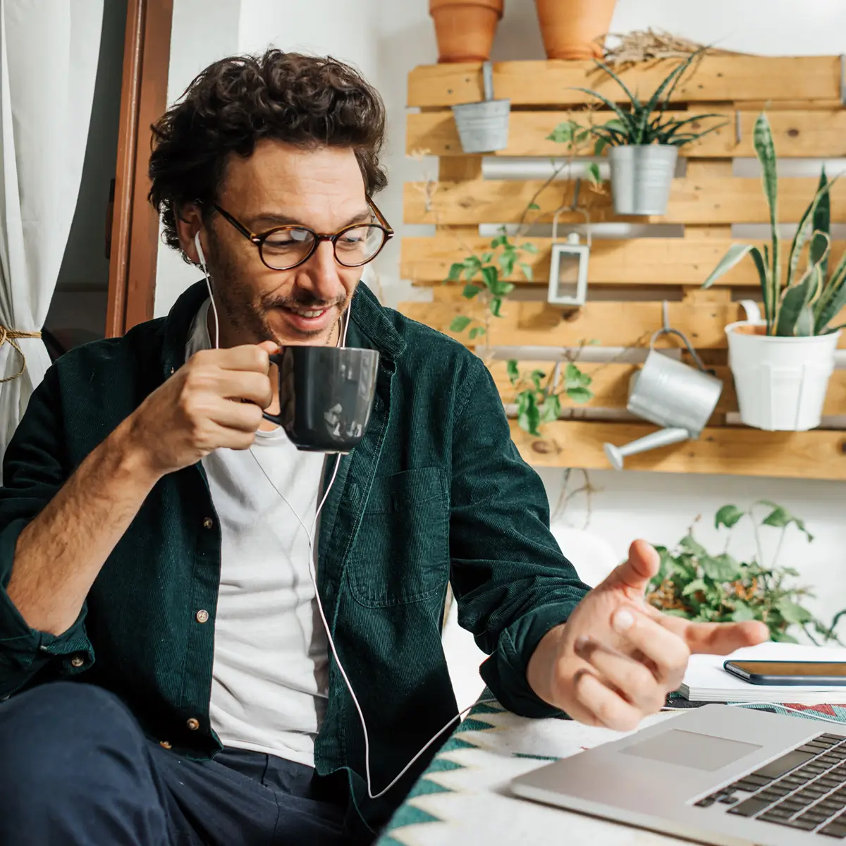 Man with glasses drinking from a black mug while using a laptop, surrounded by indoor plants and wooden wall decor.