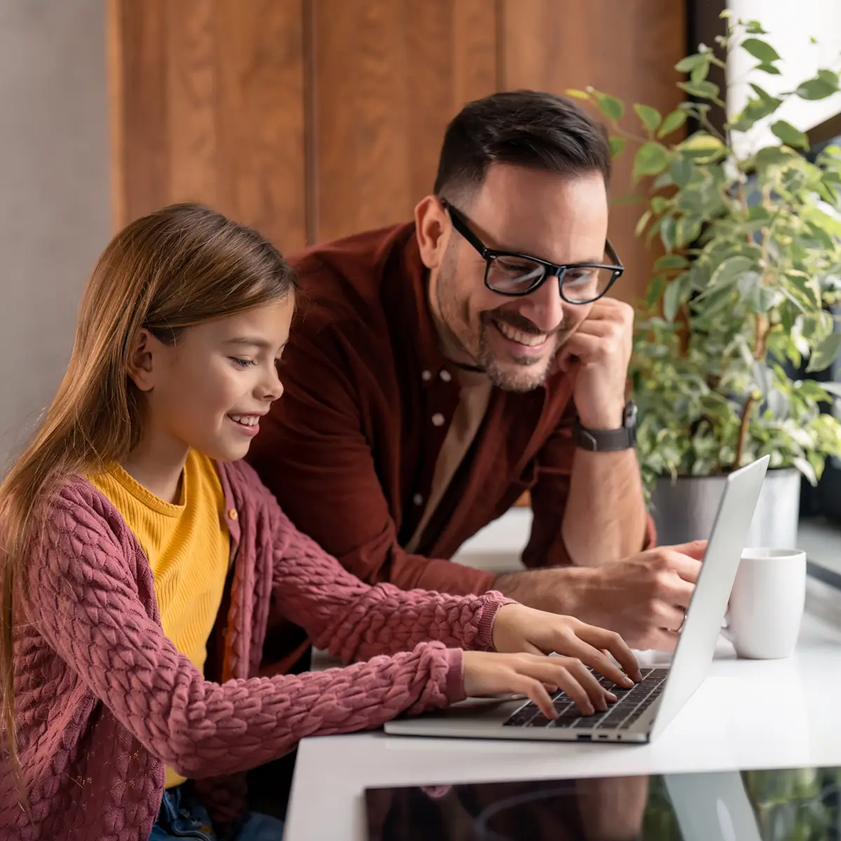 Smiling girl typing on a laptop while a man watches and encourages her at a table with a plant and coffee cup nearby.