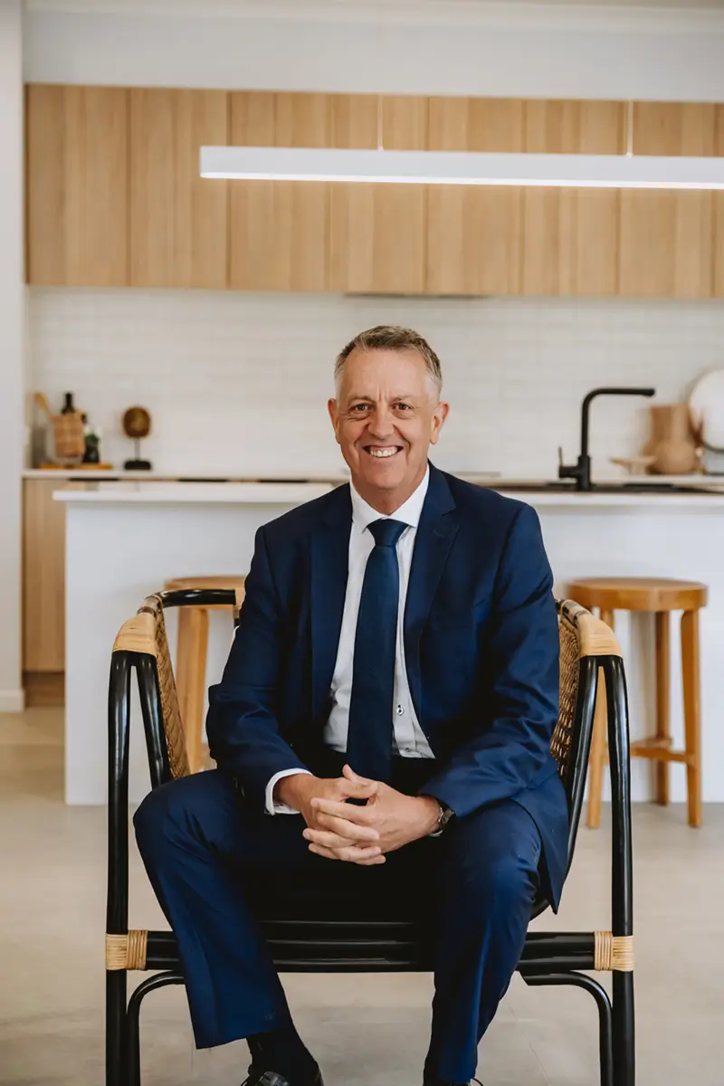 Smiling man in a blue suit sitting on a chair in a modern kitchen.