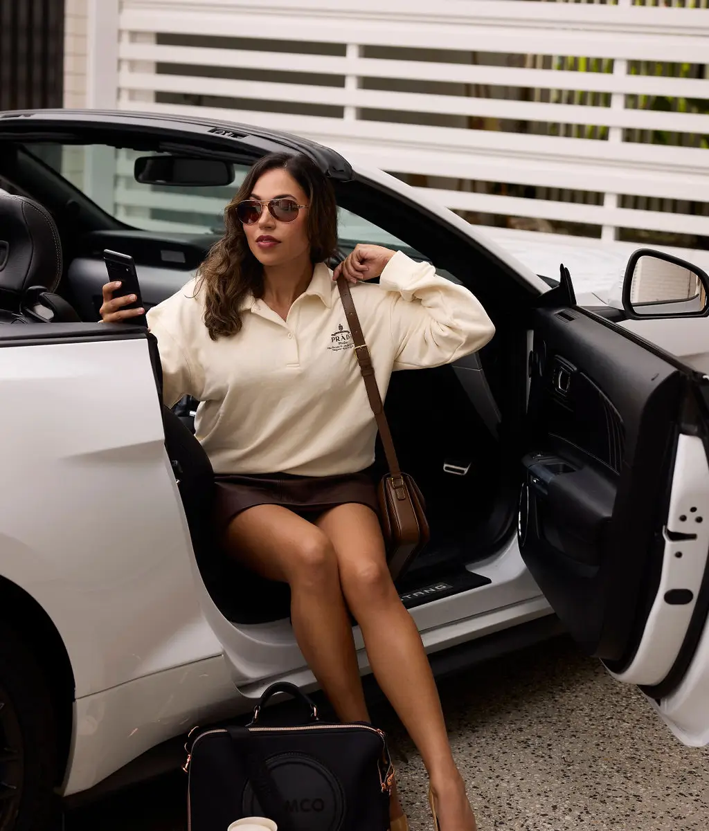 Woman wearing sunglasses and a light-colored long sleeve shirt sitting in the open door of a white car, holding a phone and a brown handbag on her shoulder.