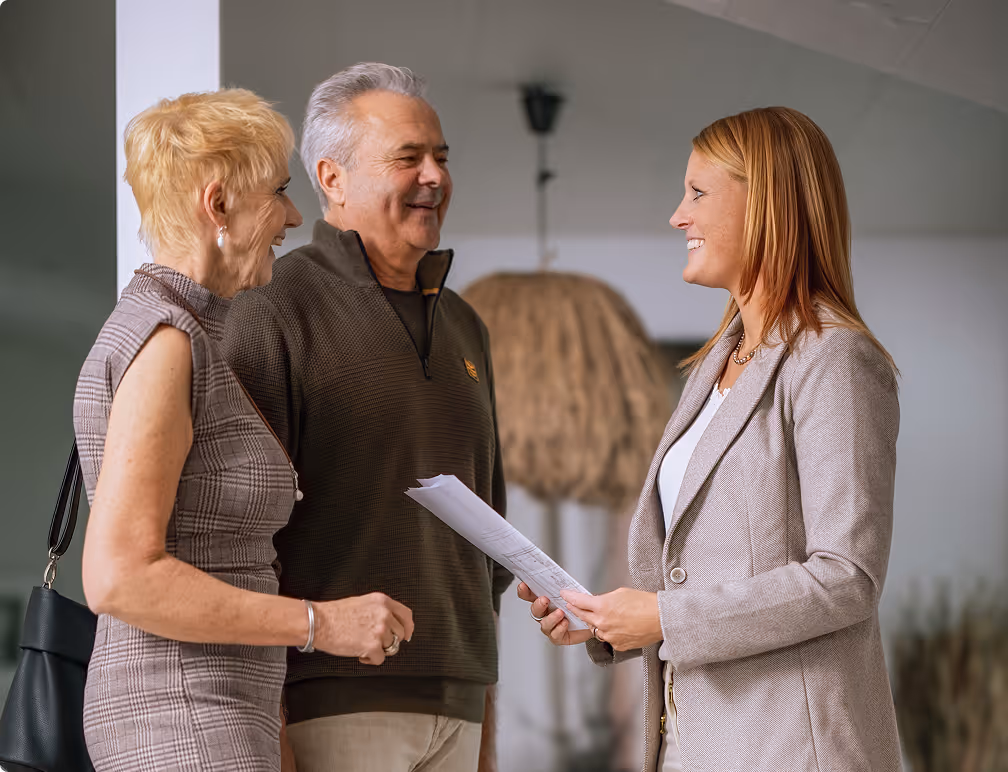 Lächelnde Frau in einem beigen Blazer, die Dokumente in der Hand hält, während sie sich mit einem Paar mittleren Alters in einem Innenraum unterhält.