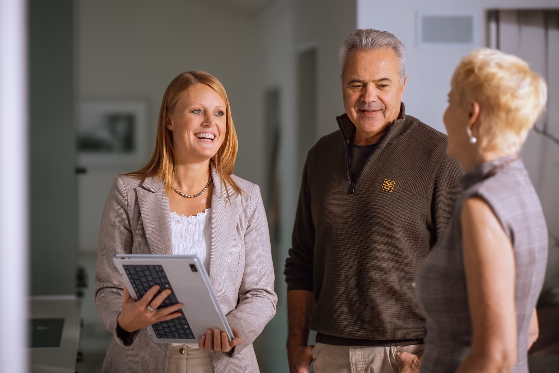 Eine lächelnde Frau mit einem Tablet spricht mit einem Mann und einer weiteren Frau in einer ungezwungenen Innenraumumgebung.