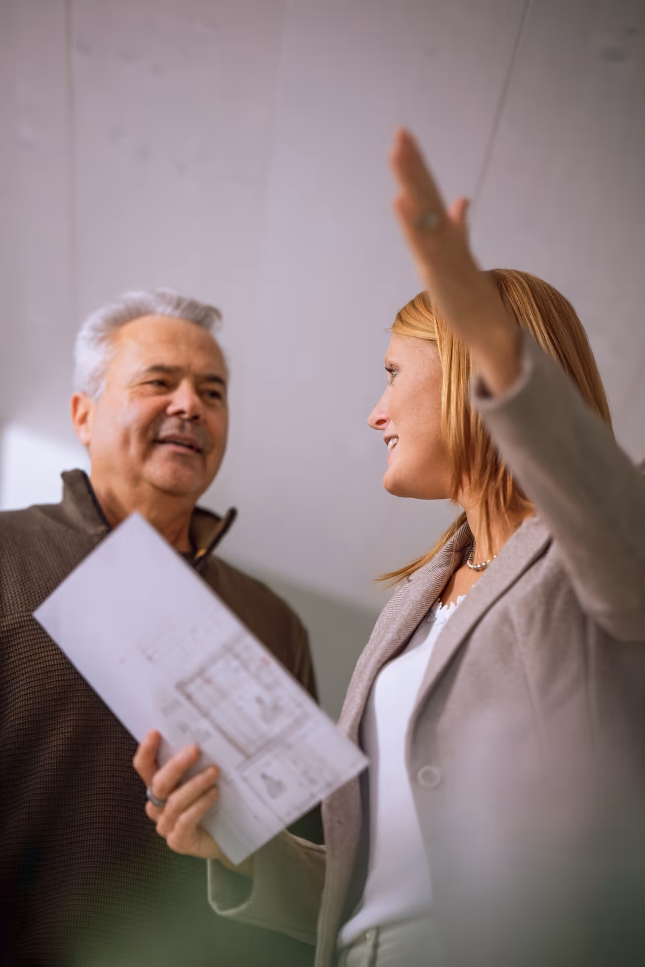 Frau, die einen Bauplan in der Hand hält und mit der Hand gestikuliert, während sie mit einem lächelnden älteren Mann spricht.
