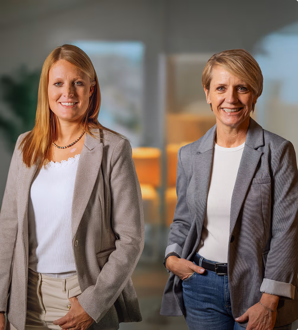 Zwei lächelnde Frauen in grauen Blazern und weißen Oberteilen, stehend vor einem unscharfen Bürohintergrund.