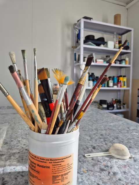Mehrere Pinsel in einem weißen Behälter auf einem marmorierten Tisch vor einem Regal mit Farben und Malutensilien.
