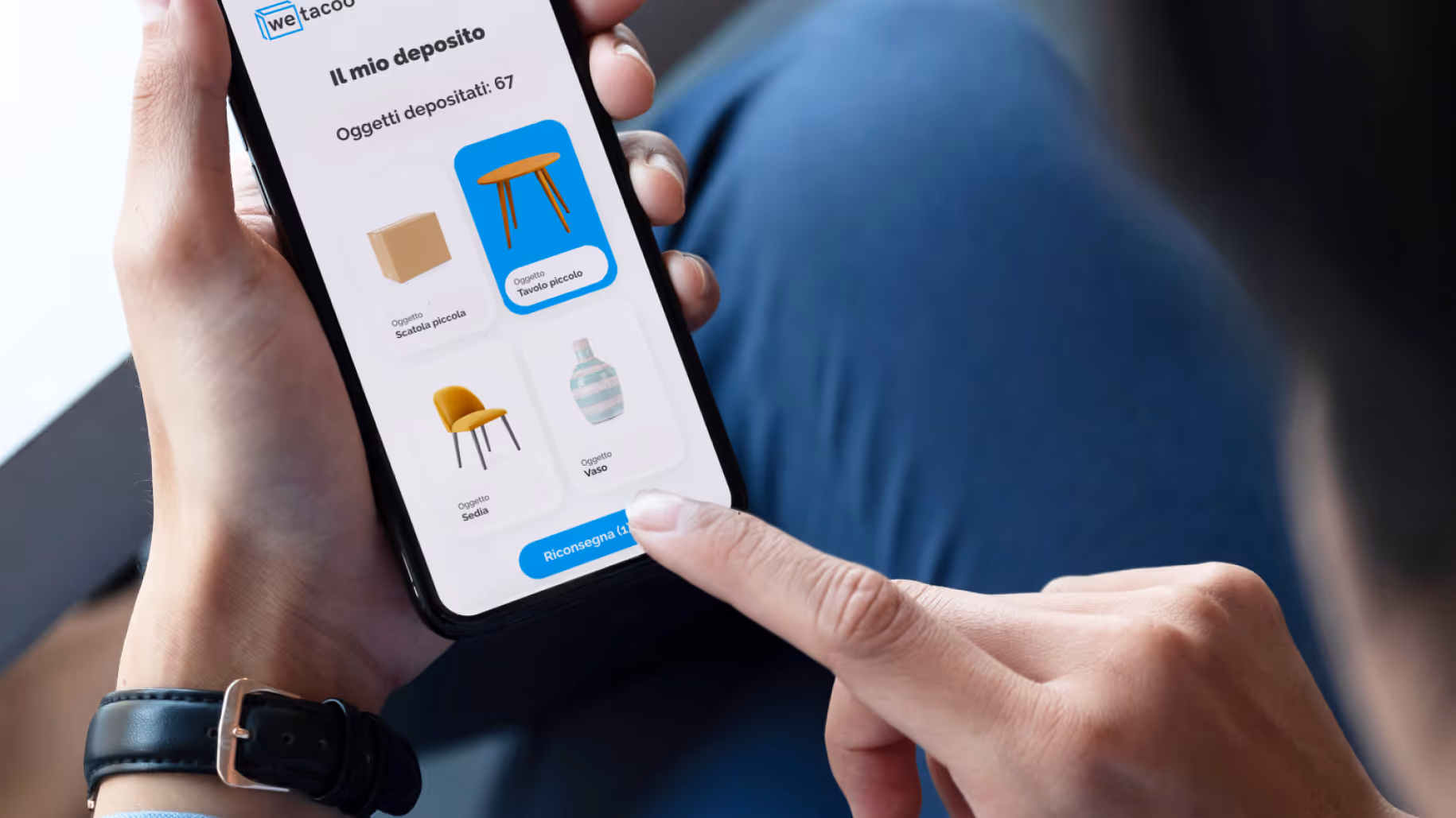 Person holding a smartphone displaying an app with a list of stored items: small box, small table, chair, and vase, with a finger about to tap the blue delivery button.