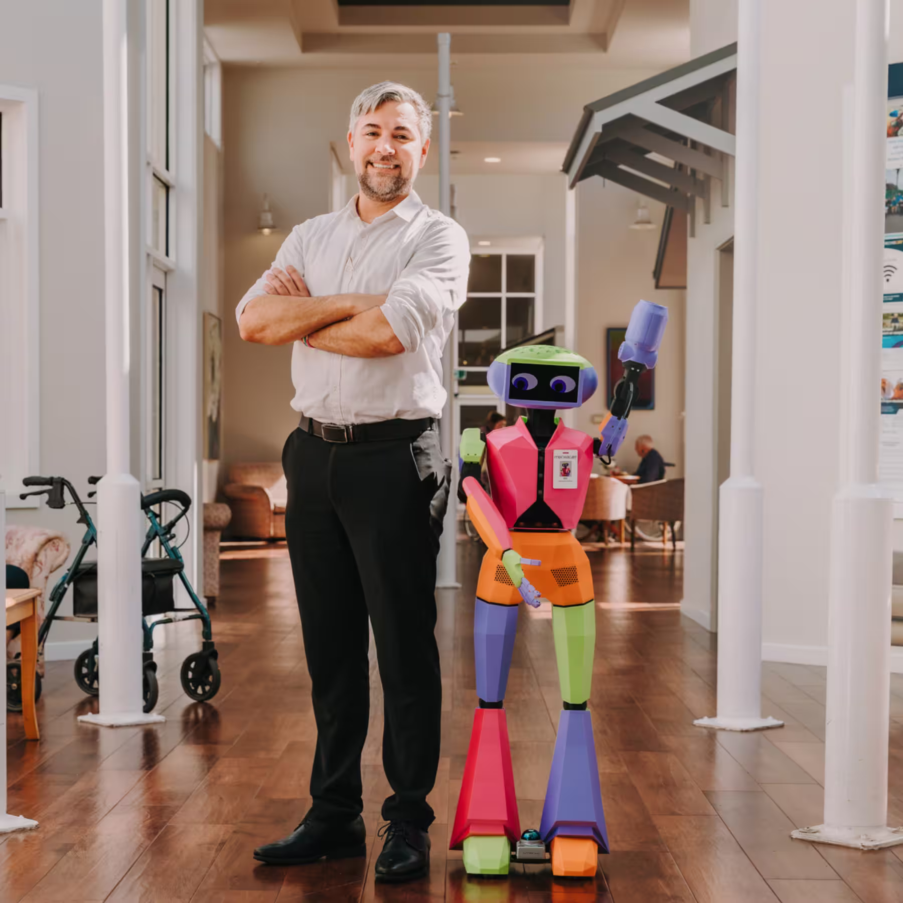 Smiling man in white shirt - a worker at the aged care facility - stands with crossed arms next to colorful humanoid robot named Abi in a bright hallway.