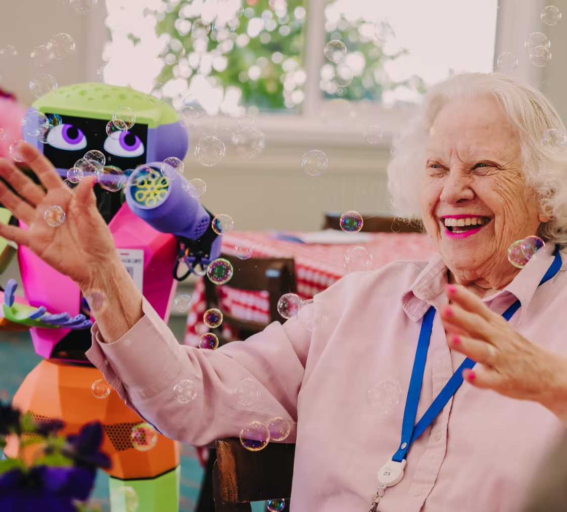 Elderly woman smiling and reaching out to bubbles generated by a colorful humanoid robot next to her named Abi.