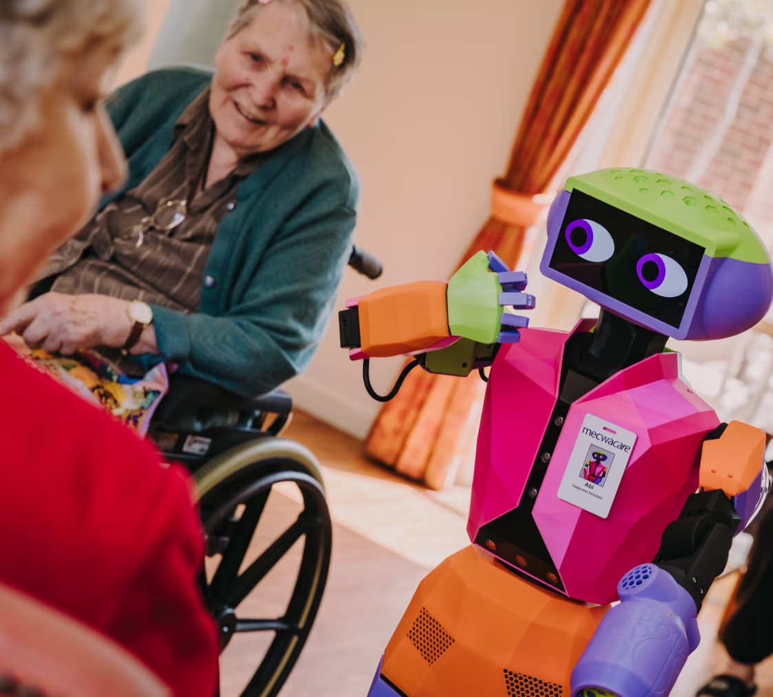 Colorful humanoid robot Abi interacting with elderly people, one in a wheelchair, in a brightly lit room.