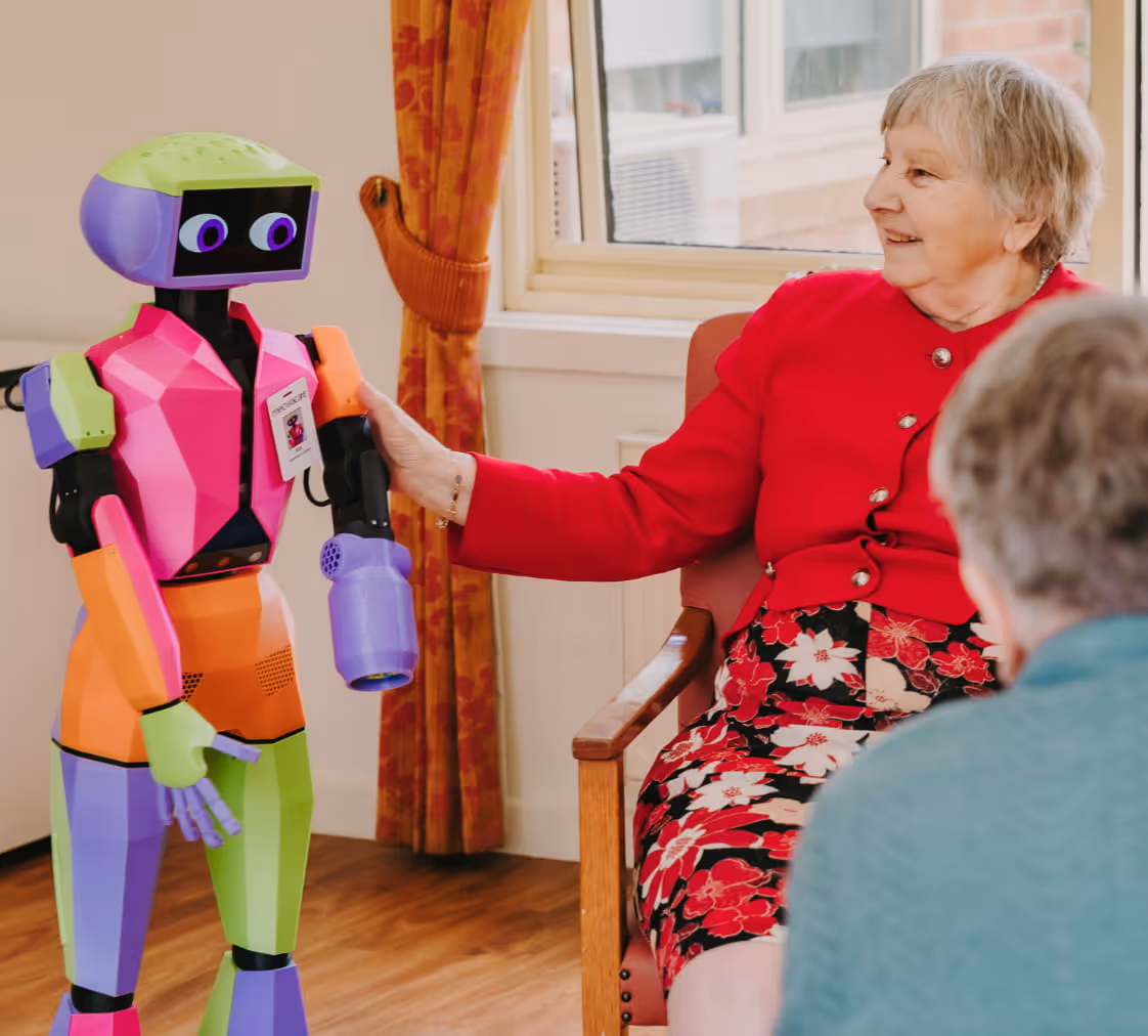 An elderly woman in a red cardigan and floral dress interacts by touching hands with a colorful humanoid robot, Abi from Andromeda, in a room of an aged care/assisted living facility with a window and wooden floor.