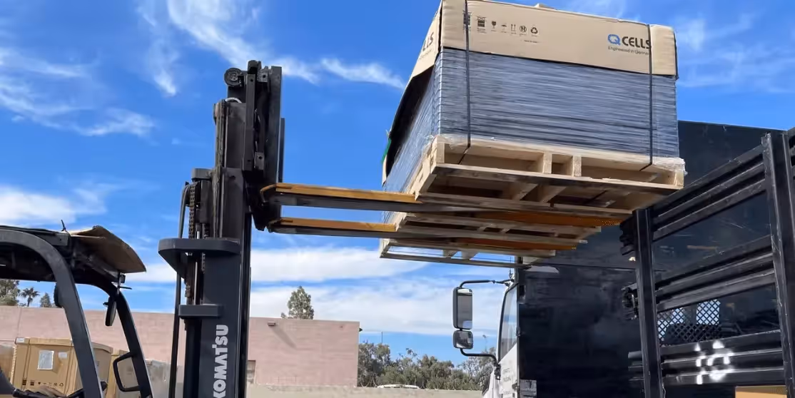 A forklift is lifting a large pallet of boxed solar panels onto a flatbed truck.