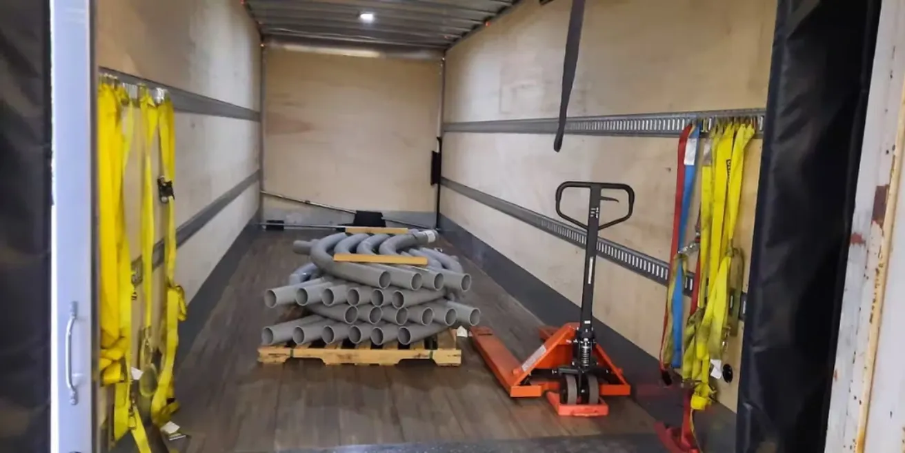 Pallet of gray conduit pipes with a pallet jack inside an LTL box truck, showing load securement straps on the wall.