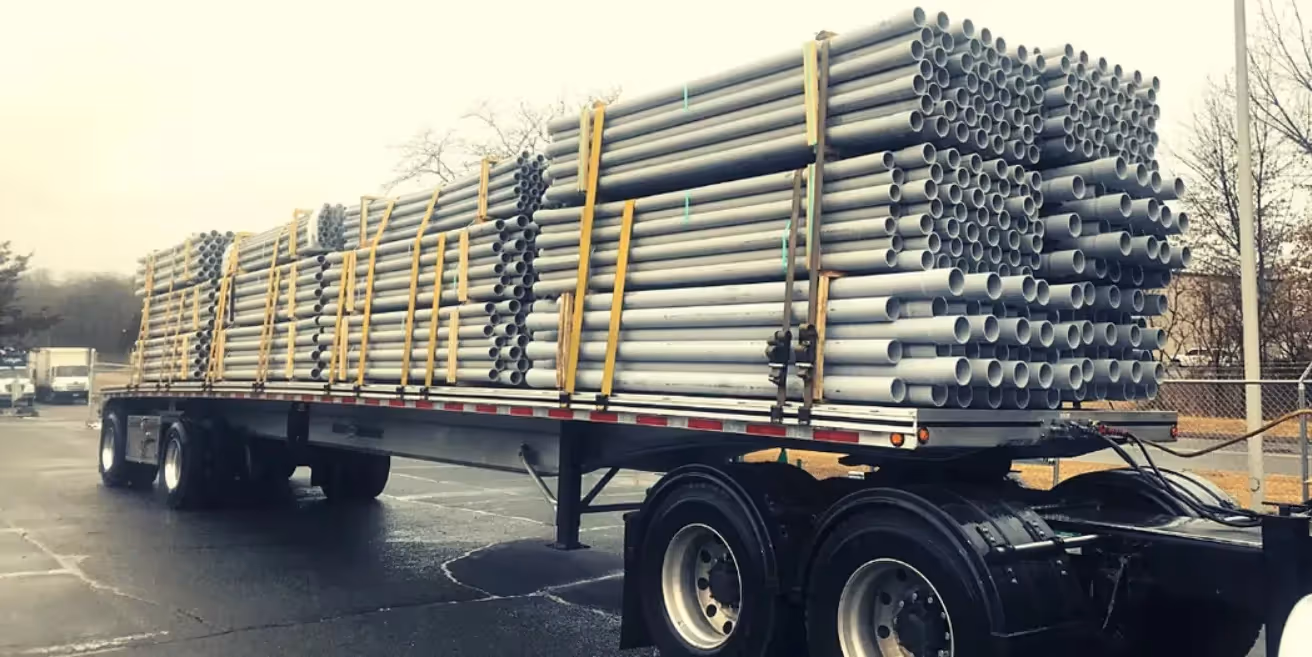 A flatbed truck carrying stacks of industrial pipes securely fastened with yellow straps parked in a lot on a cloudy day. The image highlights safe loading practices for transporting oversized and heavy equipment.