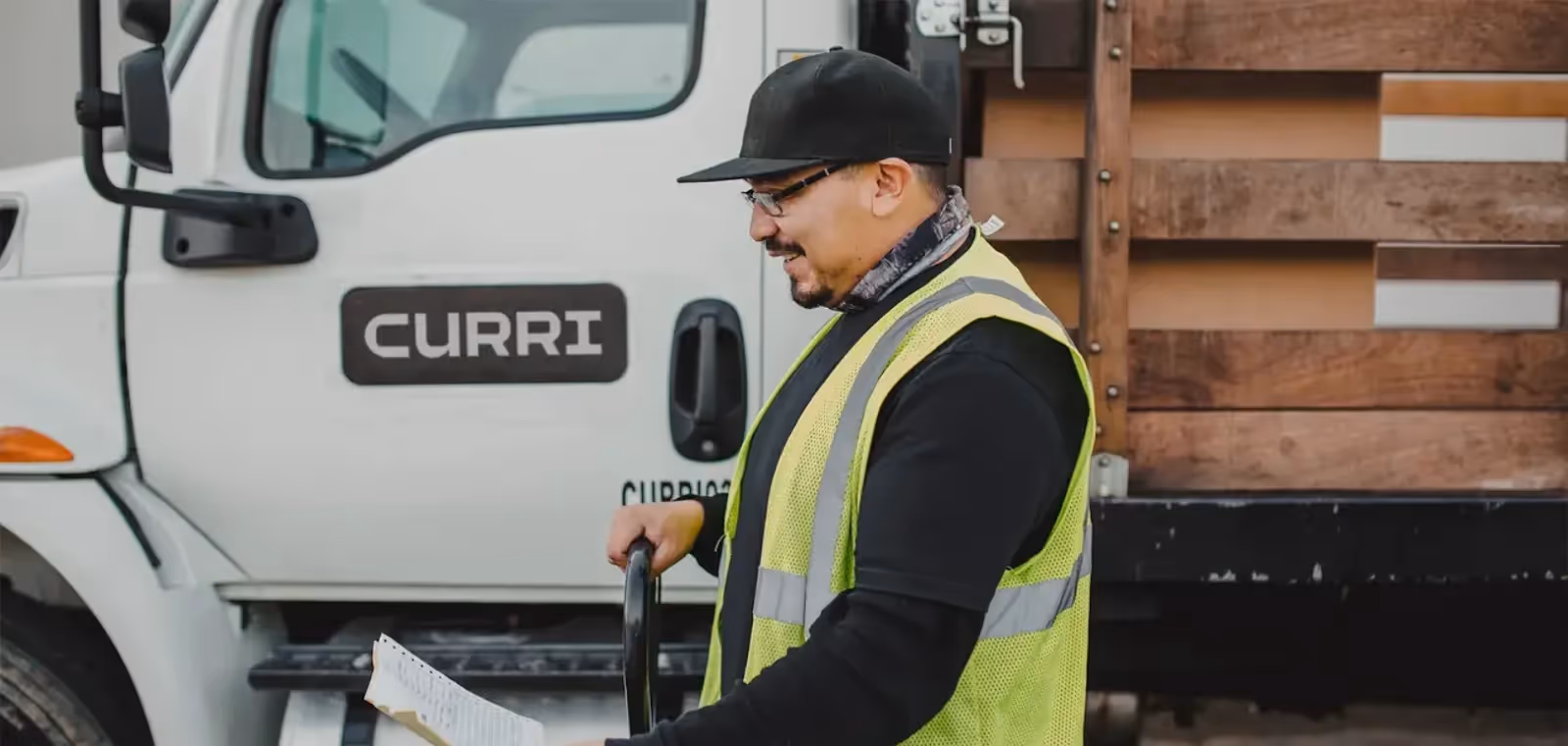 A Curri driver in a reflective safety vest reviews a delivery checklist in front of a branded Curri truck.