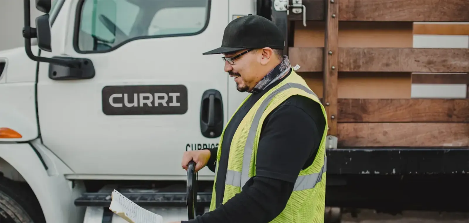 A Curri driver in a reflective safety vest reviews a delivery checklist in front of a branded Curri truck.