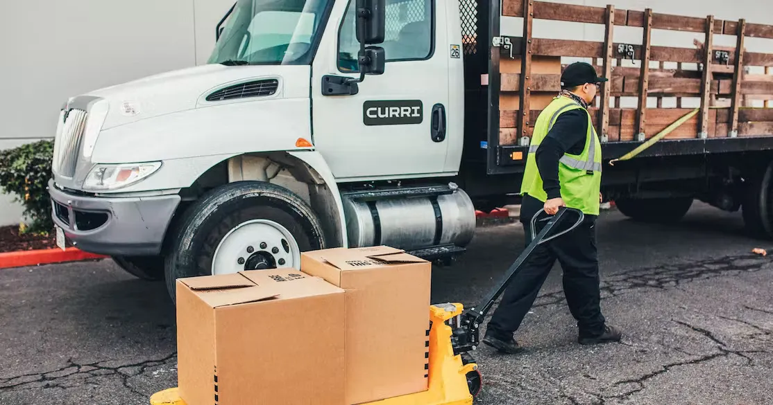 A Curri delivery truck parked beside a building while a driver in a safety vest uses a pallet jack to unload boxes, demonstrating the last-mile delivery process in action.