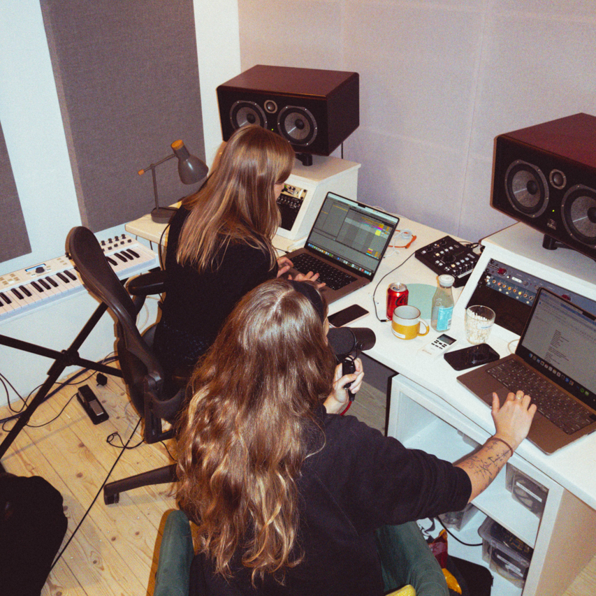 Two people working in a music studio with laptops, speakers, a microphone, and a keyboard on a wooden floor.
