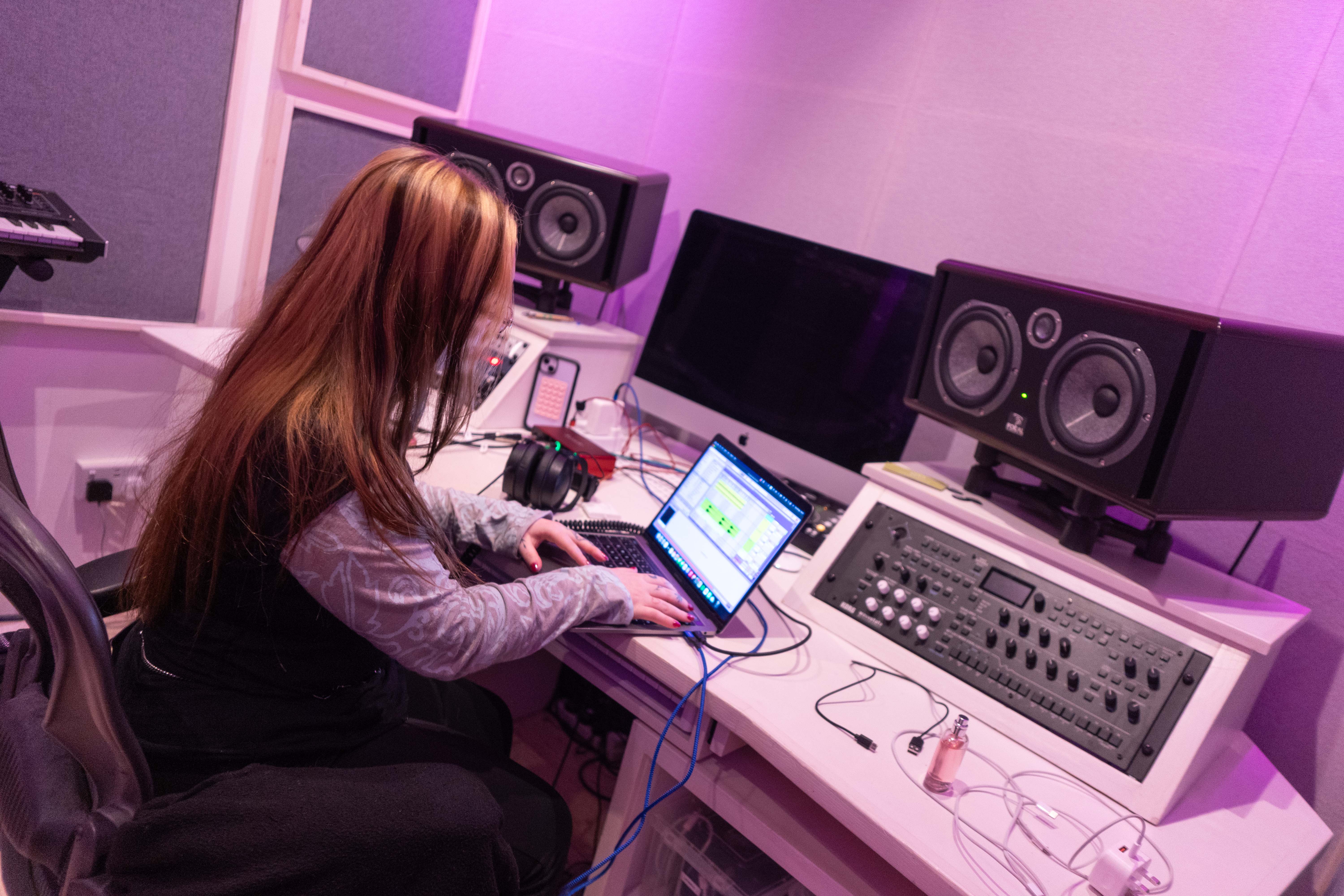 Person working on a laptop in a music studio with large speakers, audio equipment, and purple lighting.