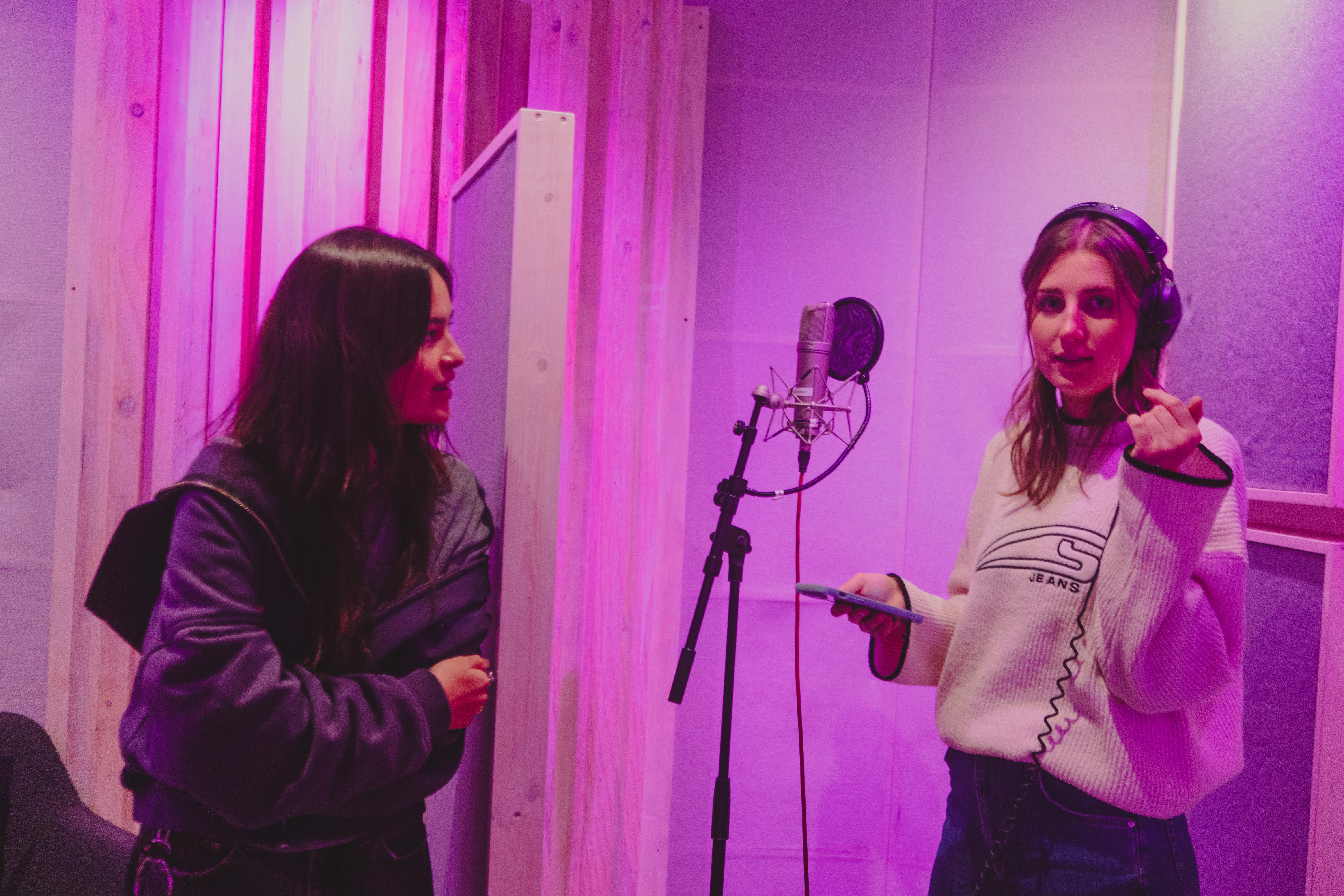 Two women in a recording studio, one wearing headphones and holding a phone near a microphone, the other looking at her, both under purple lighting.