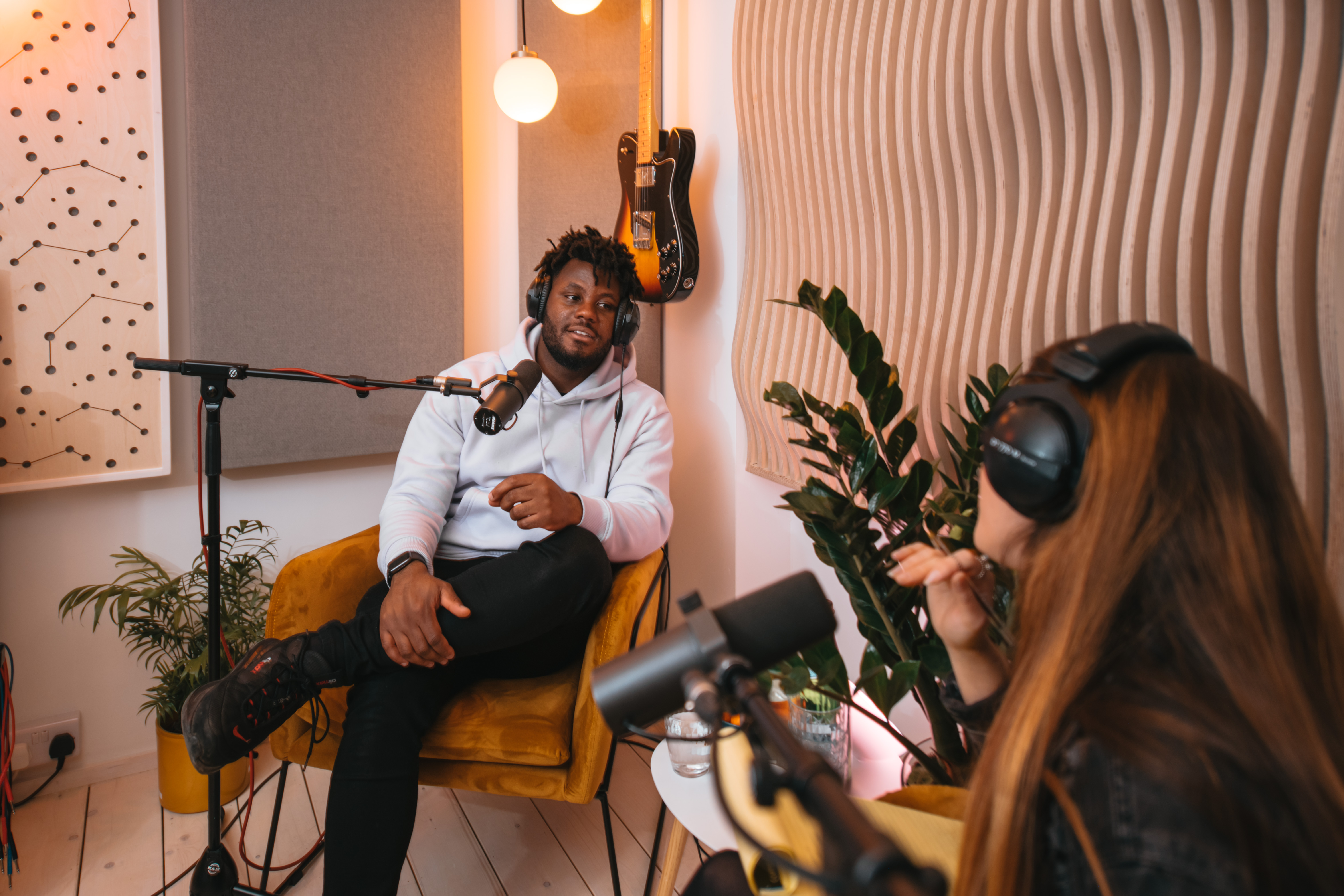 Two people recording a podcast in a modern studio, wearing headphones and speaking into microphones with guitars and plants in the background.