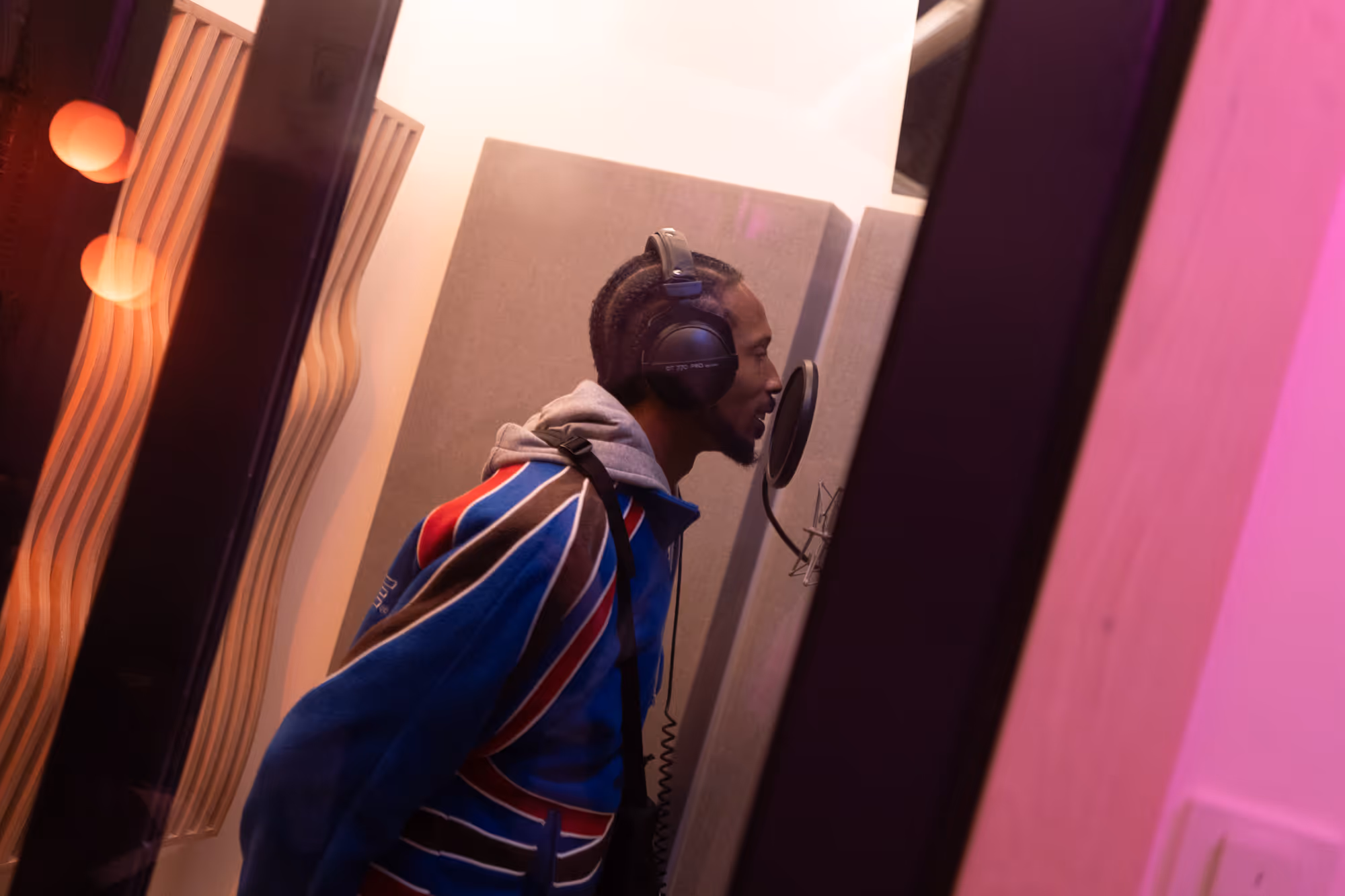 Rap artist recording vocals in Studio B at Noatune Studios, Hackney, using a Neumann U87 microphone inside a soundproof vocal booth with warm lighting and wooden acoustic diffusers.