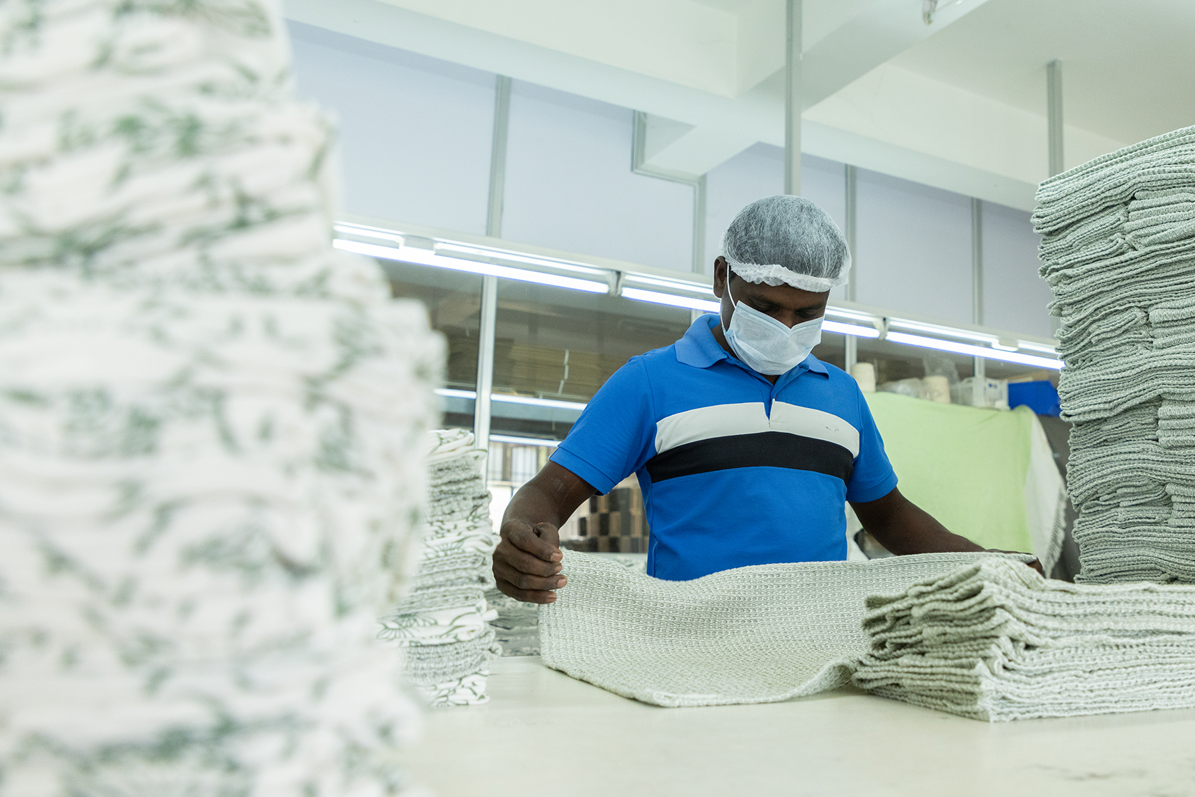 Factory worker folding a knitted textile on a table surrounded by stacks of similar textiles.