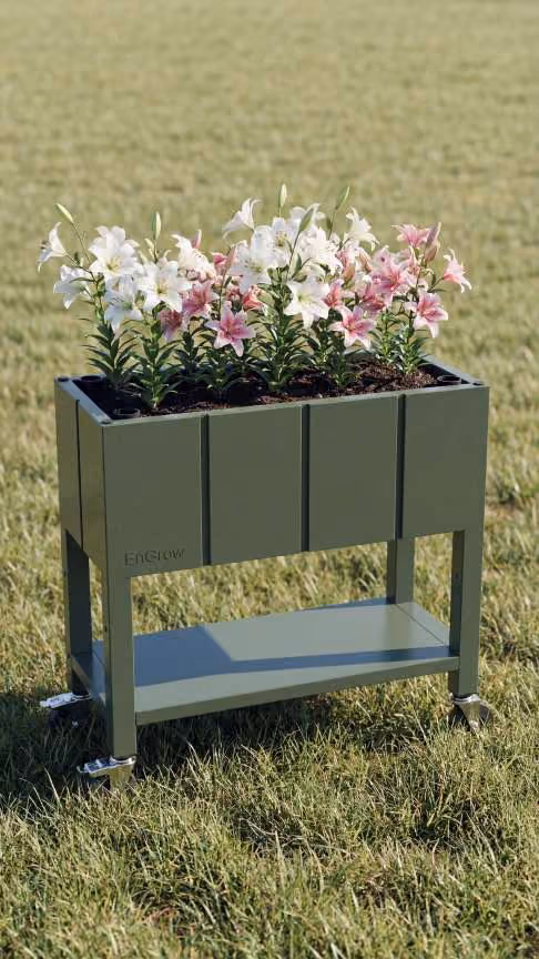 The Garden Bed in green color on wheels with shelf, filled with blooming white and pink lilies, standing on grassy field.