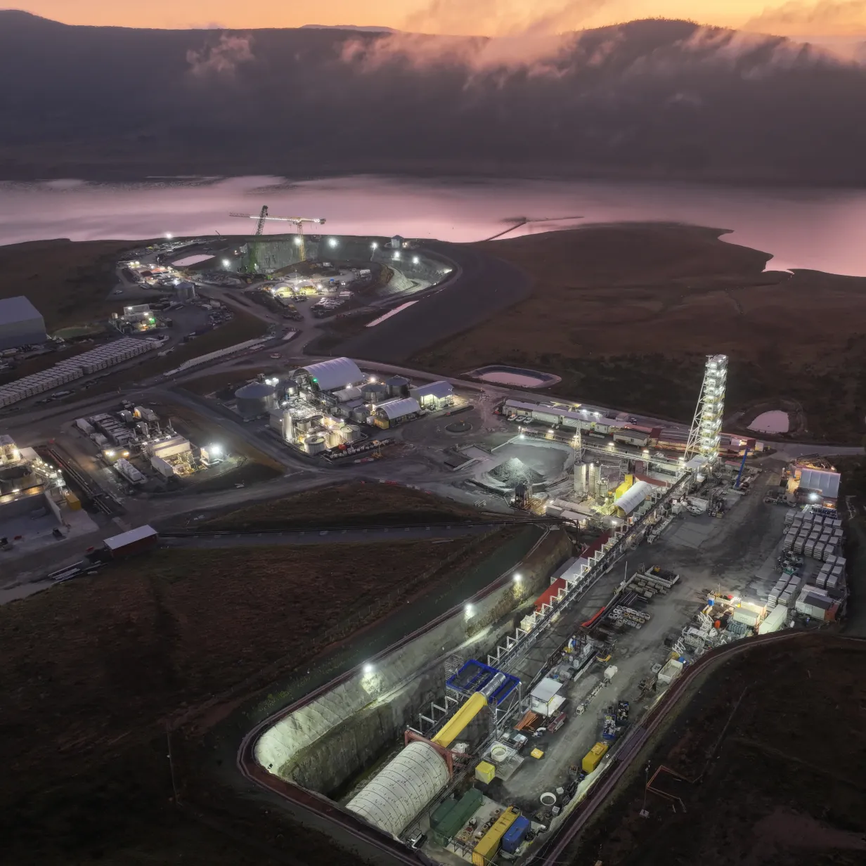 Aerial view of a large industrial construction site at dusk with illuminated buildings and machinery surrounded by a river and hills.