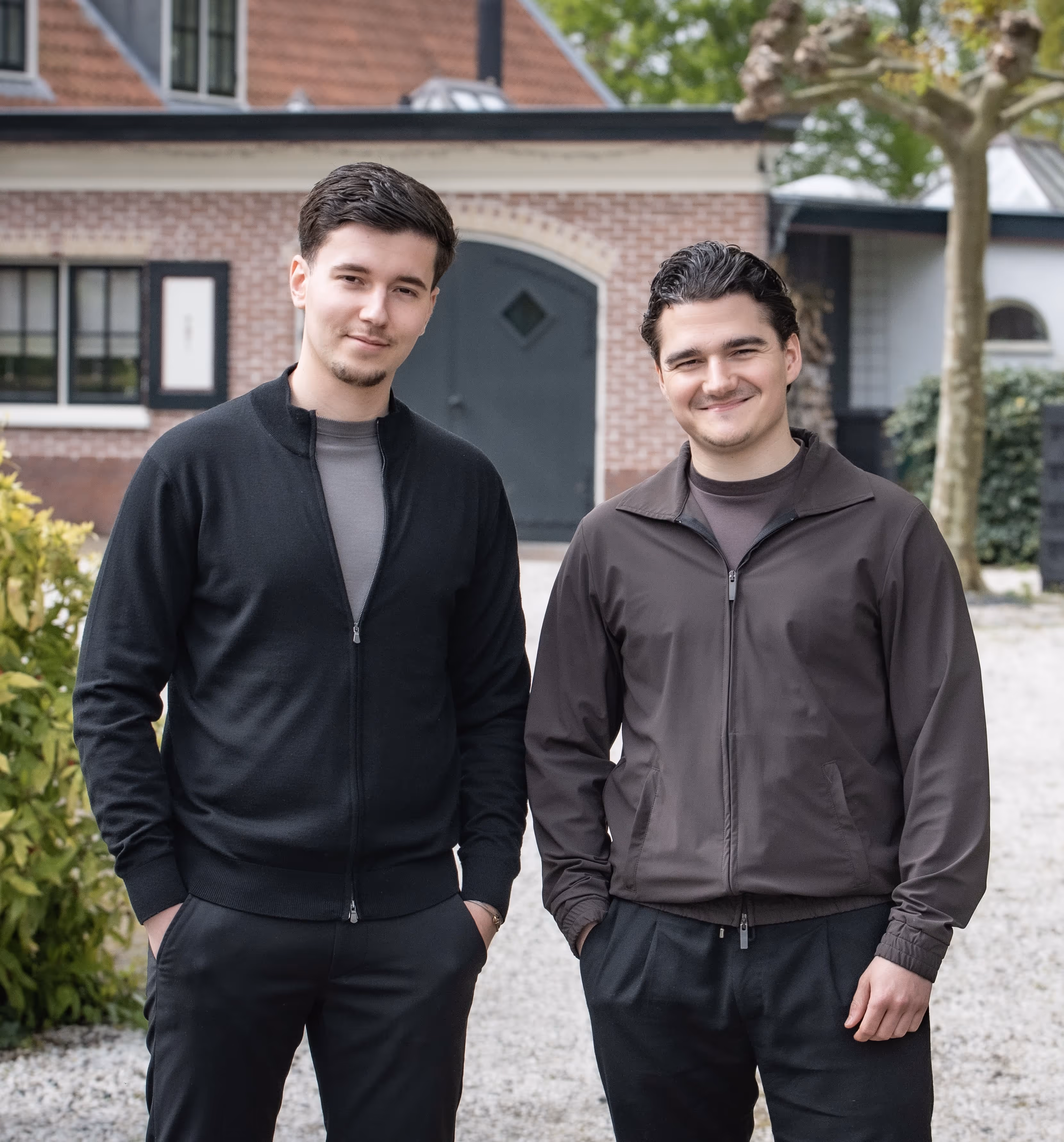 Two young men standing outdoors in casual jackets, smiling in front of a brick building with windows and a black door.