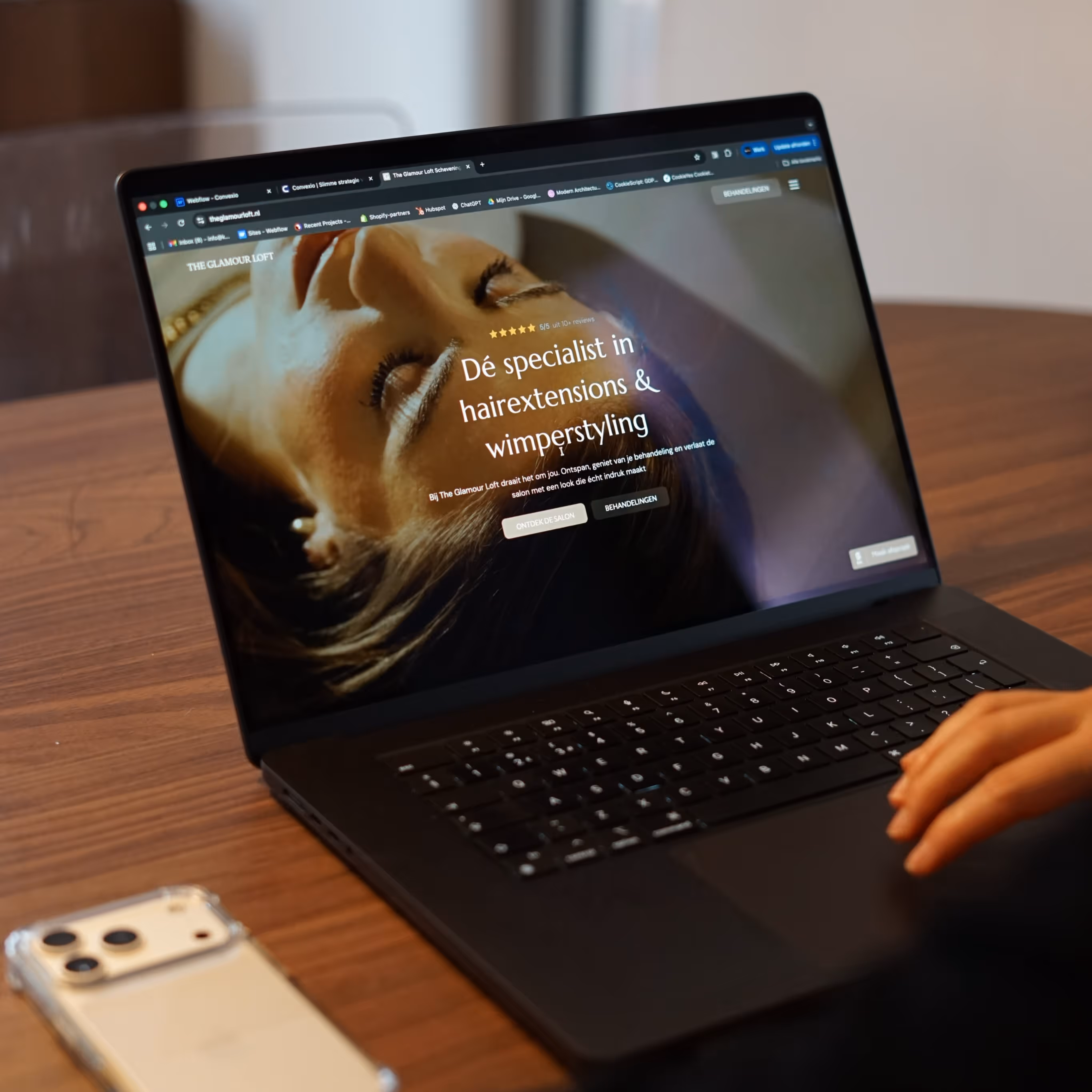 Person's hand using a laptop displaying a beauty salon website specializing in hair extensions and eyelash styling.