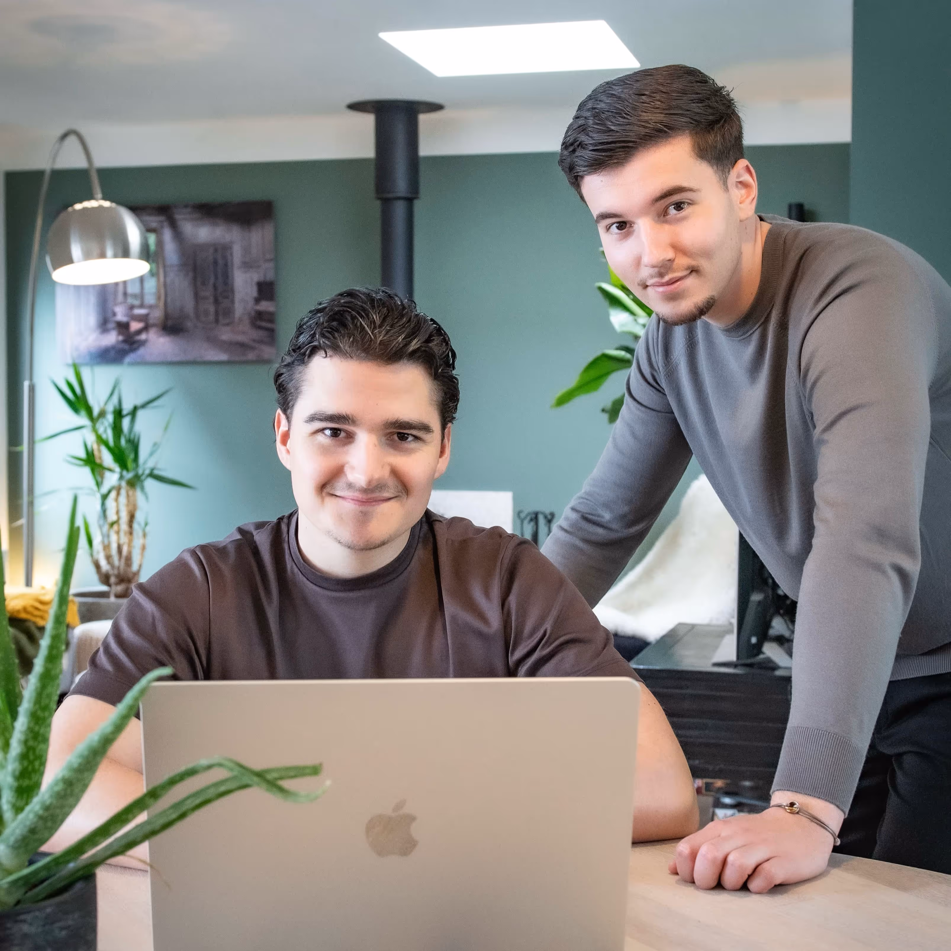 Two young men smiling, one seated behind an open laptop and the other leaning on the table in a modern room with plants and green walls.