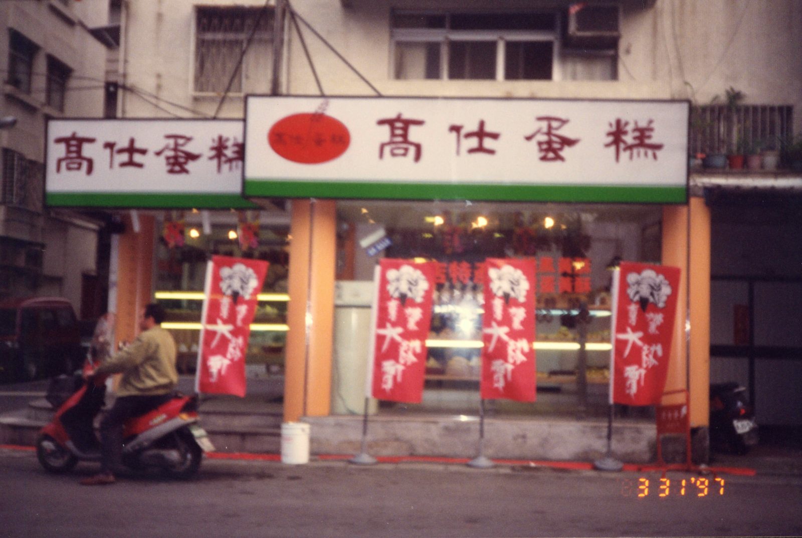 Kao Shieh Bakery’s storefront by day in 1997: white signs with red lettering, orange pillars, red banners and a man on a red scooter