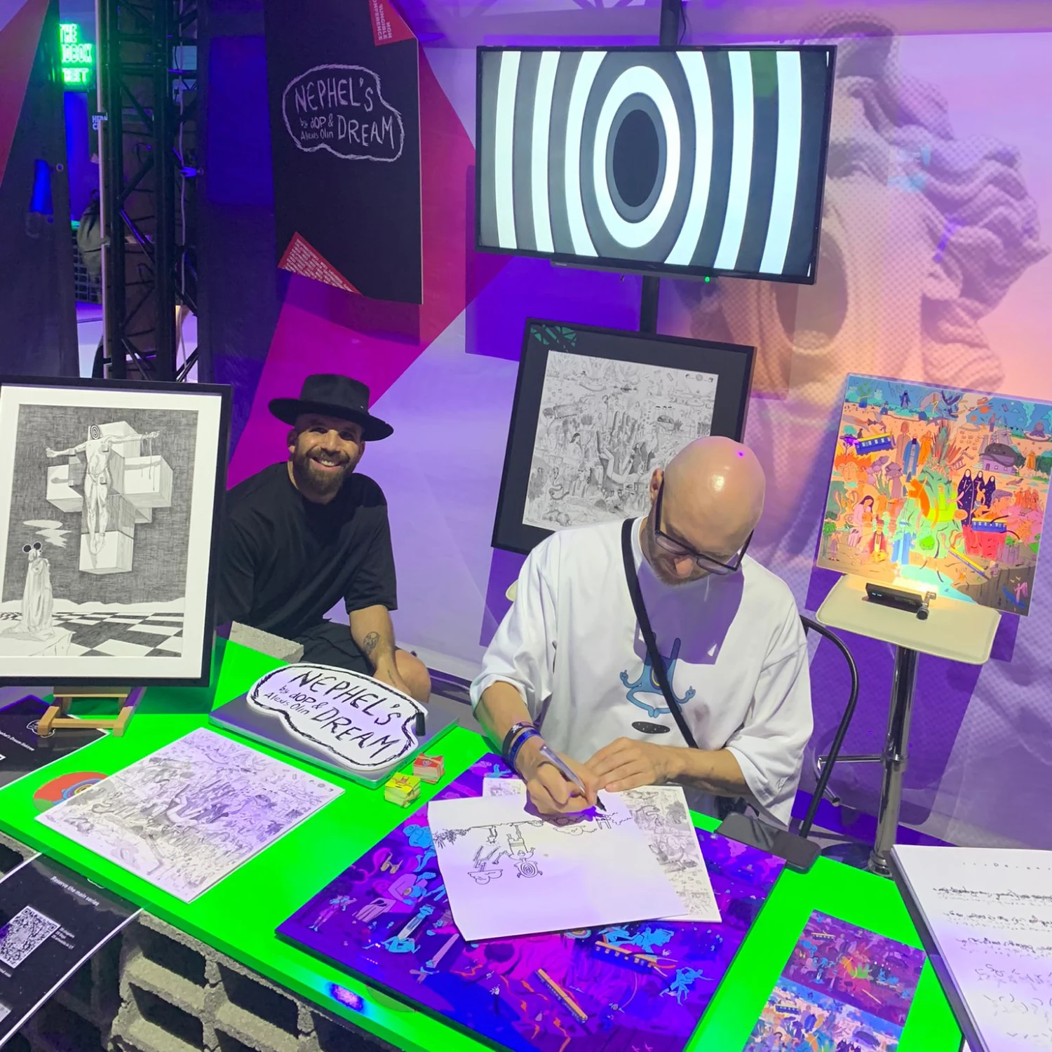 Nephel’s Dream exhibition stand with artworks, a screen and an artist drawing at a table.