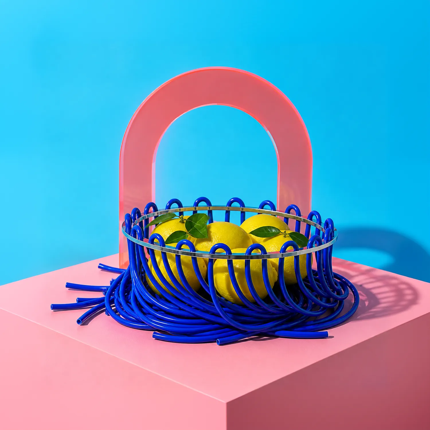 Lace fruit bowl with blue rubber cords and lemons, on a pink surface against a cyan background.