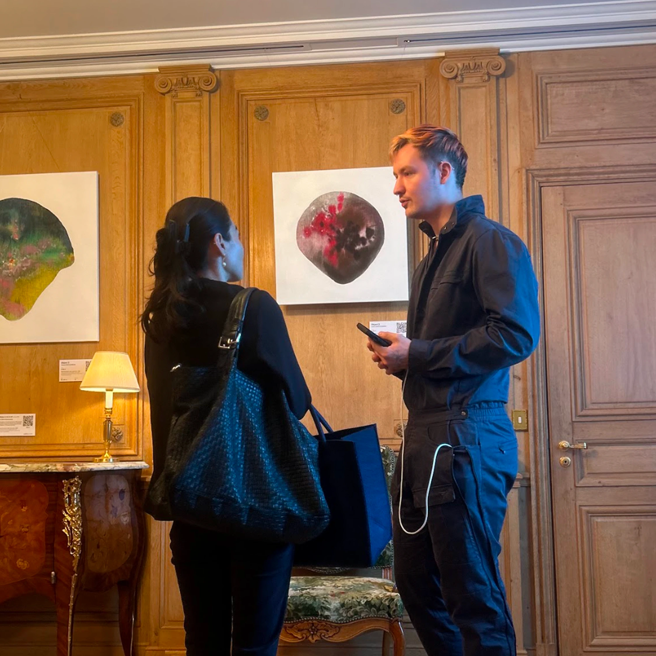 Two visitors talking in front of paintings on a wooden gallery wall.