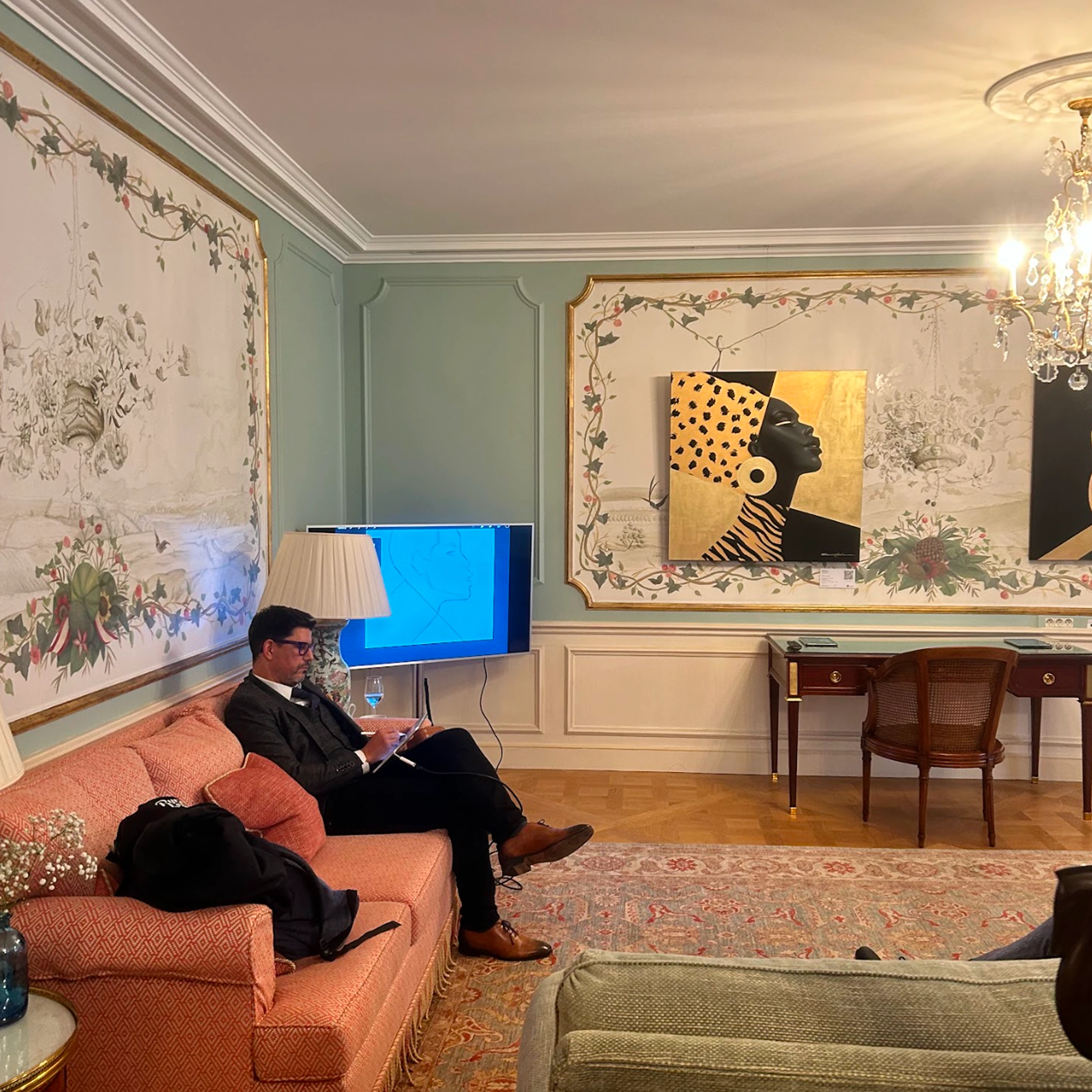 A seated visitor in a decorated salon with a portrait on the wall.