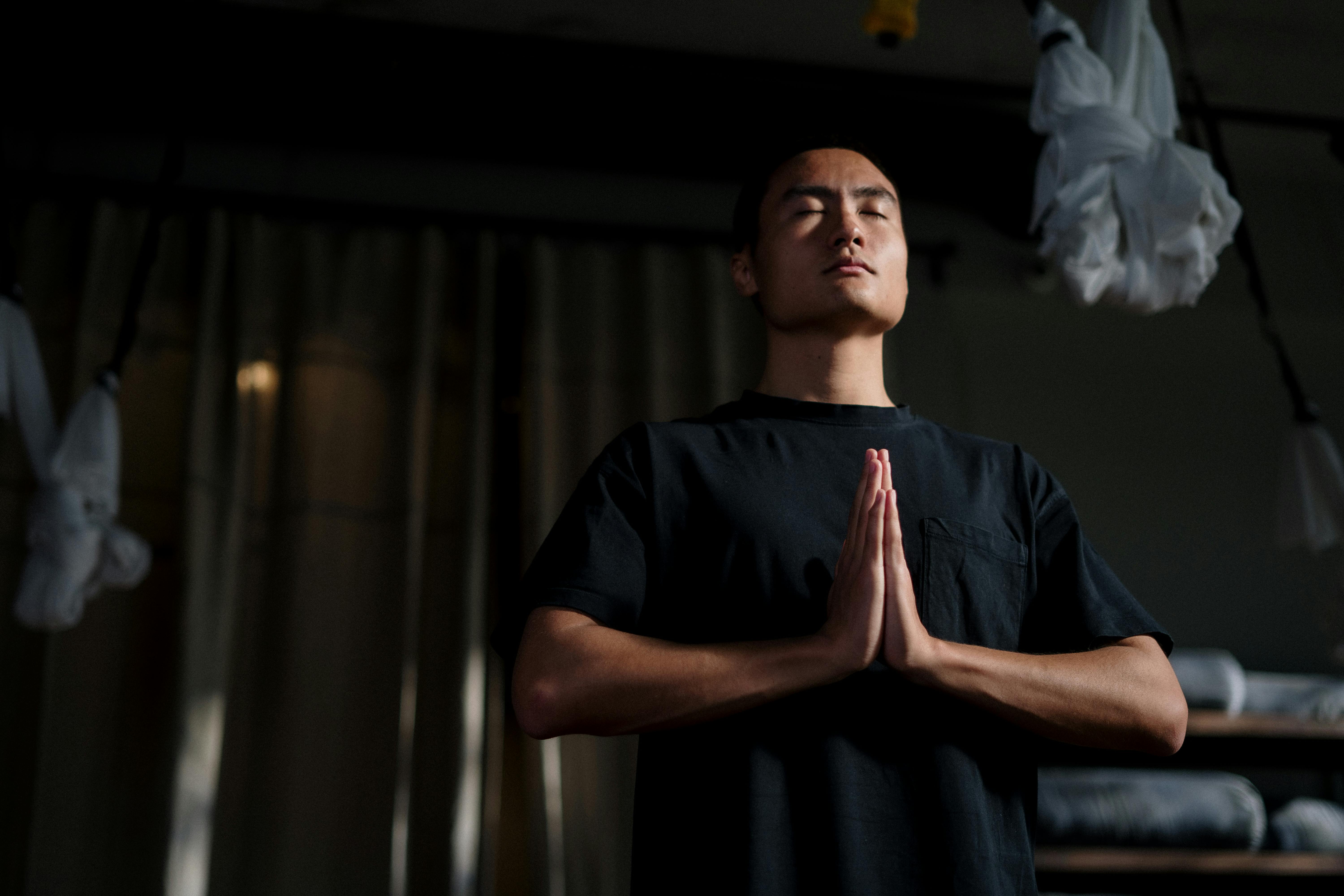 Man with eyes closed and hands in prayer position, meditating indoors with curtains and hanging fabric in background.
