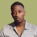 Portrait of a serious young Black man with short hair and a trimmed beard wearing a gray plaid shirt against a light green background.