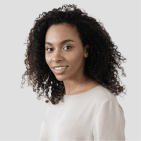 Portrait of a smiling young woman with curly black hair wearing a light beige top against a light gray background.