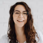 Smiling woman with long curly brown hair and glasses against a white brick wall.