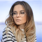 Young woman with long ombre hair and round glasses wearing a striped top looking to the side with a blurred blue sky background.