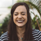 Smiling woman with closed eyes and dark lipstick wearing a striped shirt and necklace in an outdoor setting.