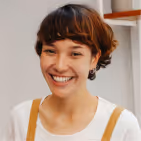 Smiling young person with short brown hair and white shirt, standing indoors near a wall corner.