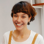Smiling young person with short brown hair and white shirt, standing indoors near a wall corner.